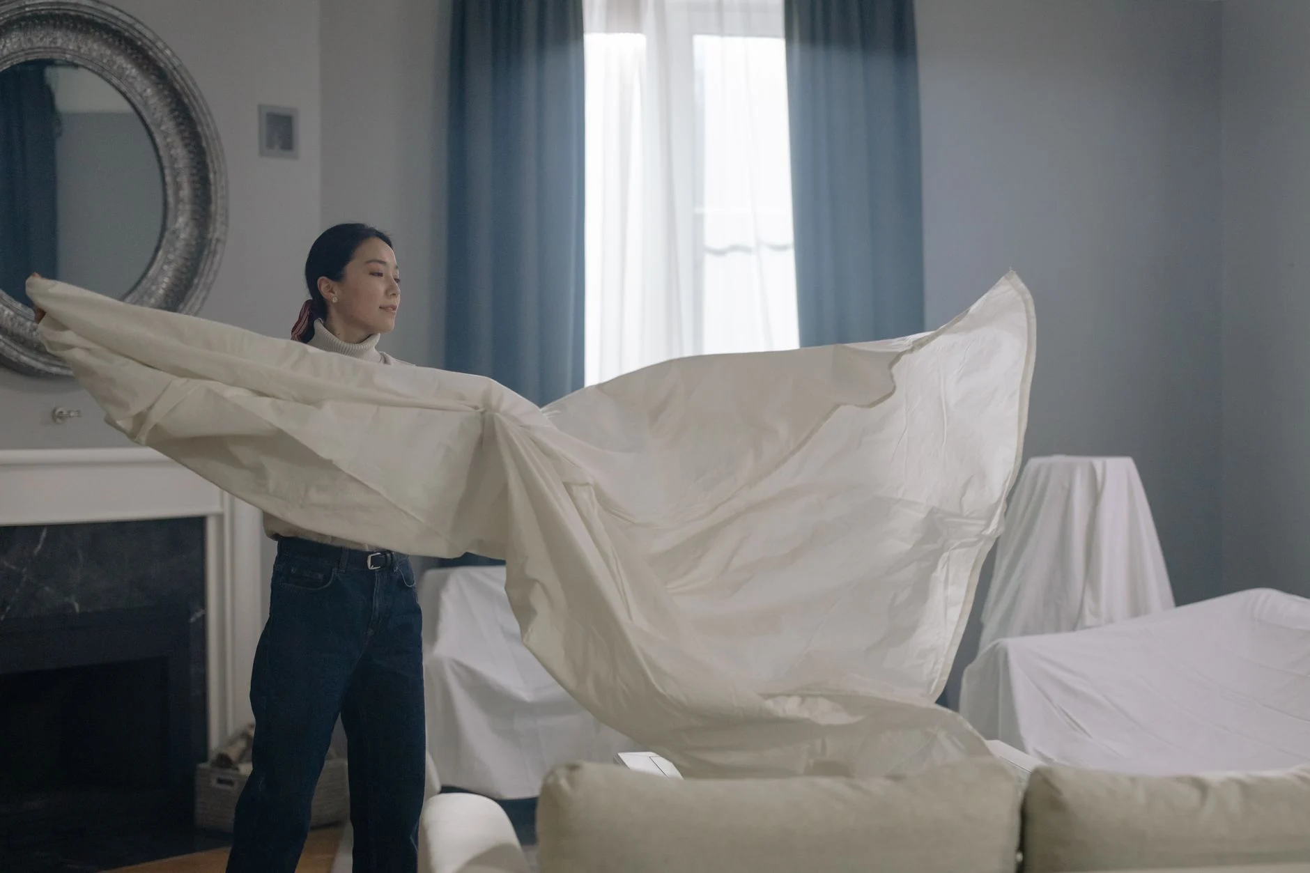 Asian woman in elegant room fluffing a large white sheet, showcasing interior decor.