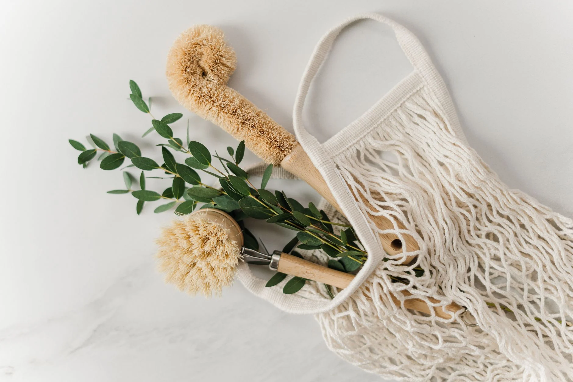 Eco-friendly cleaning brushes and eucalyptus branch in a net bag on a marble surface.