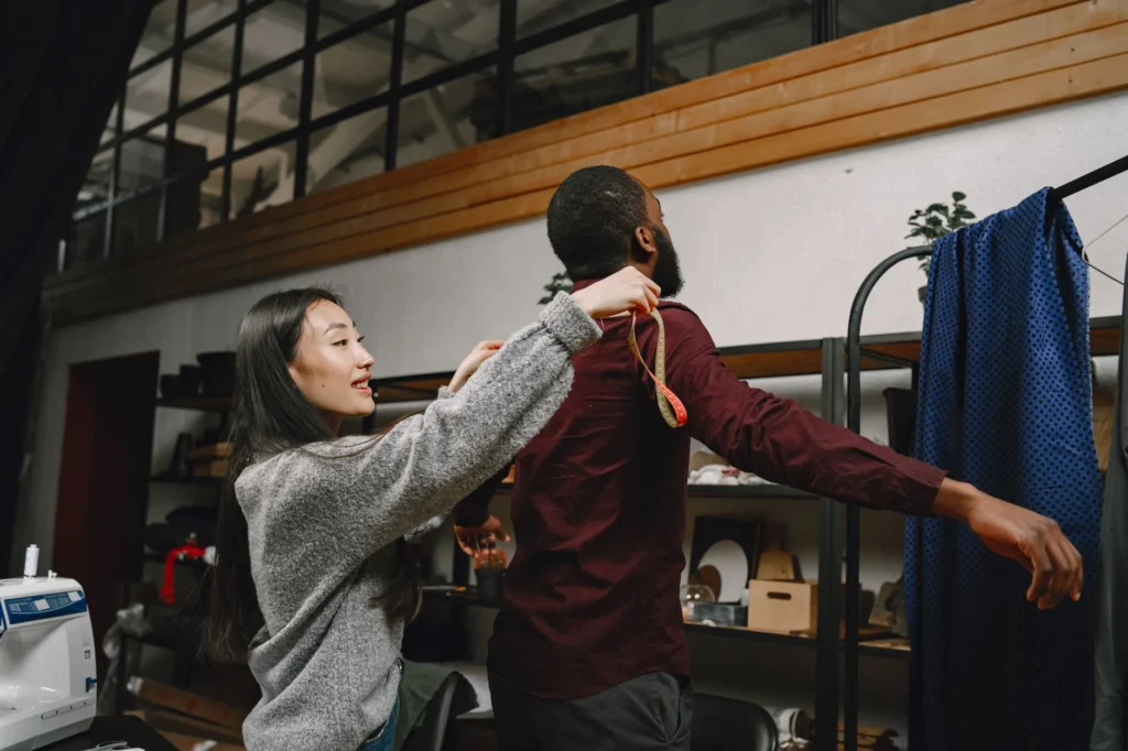 Seamstress using tape to measure a client's arm in a workshop setting, illustrating tailoring and craft.