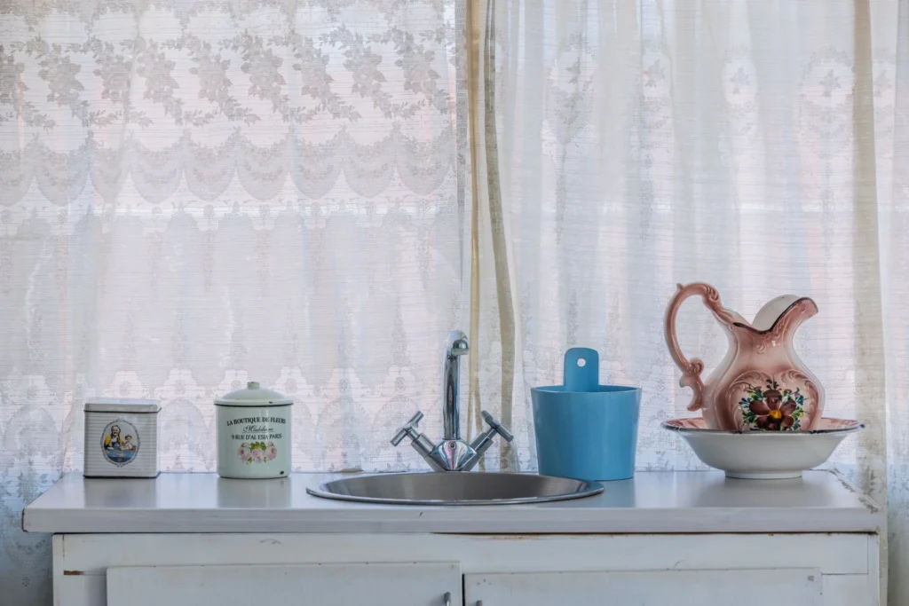 A vintage kitchen scene with decorative jars, a floral pitcher, and lace curtains.