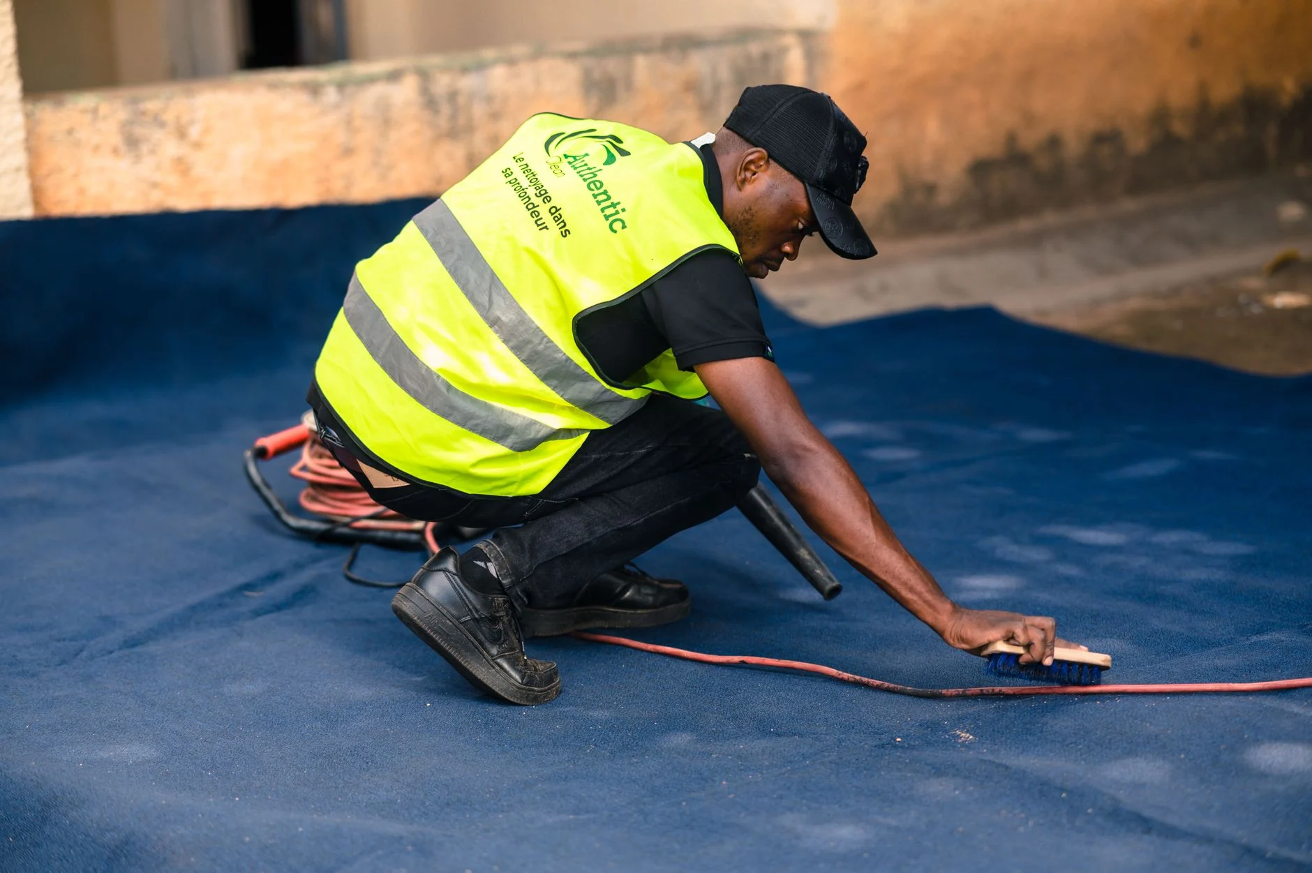 A worker dressed in a safety vest cleans a blue carpet outdoors with a brush.