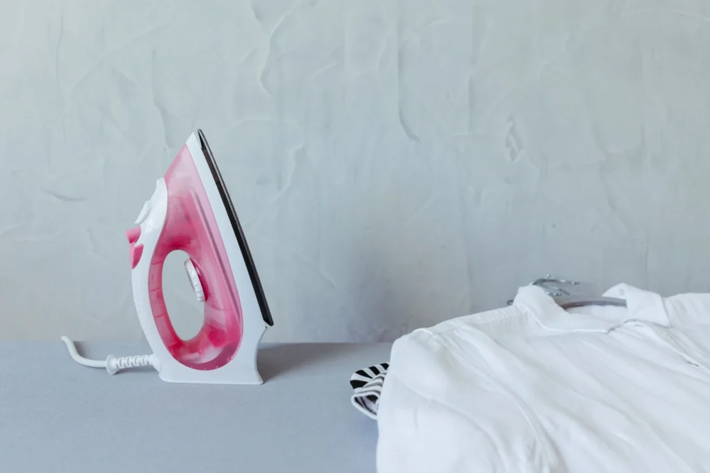 A vibrant pink iron next to a neatly folded white shirt on an ironing board against a textured wall.