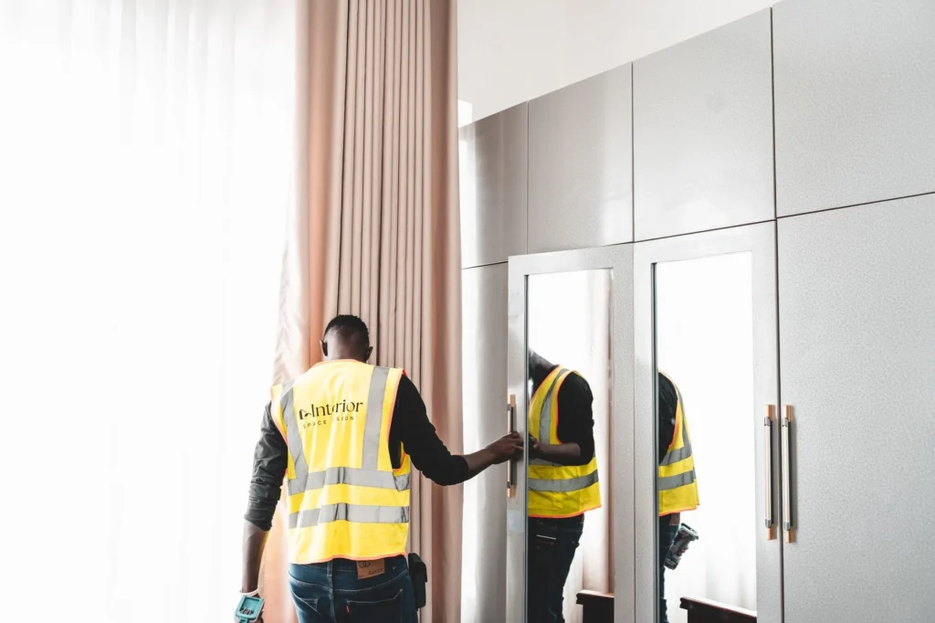 A maintenance worker in a safety vest examines a wardrobe mirror indoors.