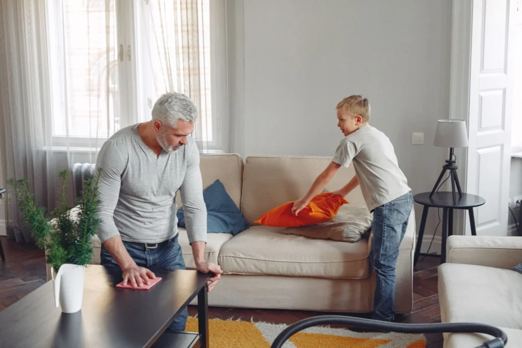 A father and son clean their living room, tidying up cushions and dusting tables.