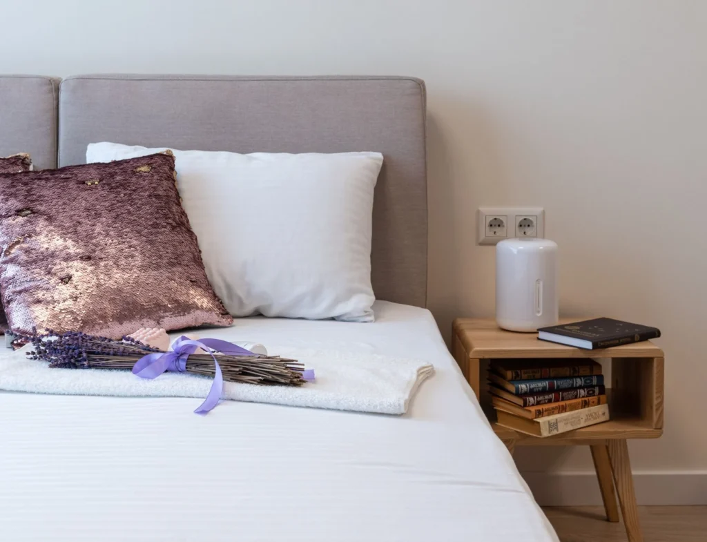 Bunch of fresh aromatic lavender placed on comfortable bed with decorative pillows near wooden night table with stack of books