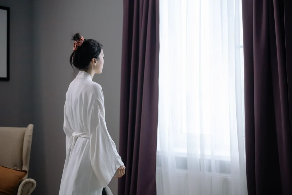 Woman in a white robe standing indoors looking out a window with sheer curtains.