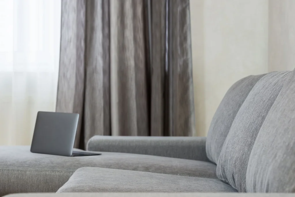 Modern flat interior with open netbook placed on couch with pillows against window with gray curtains