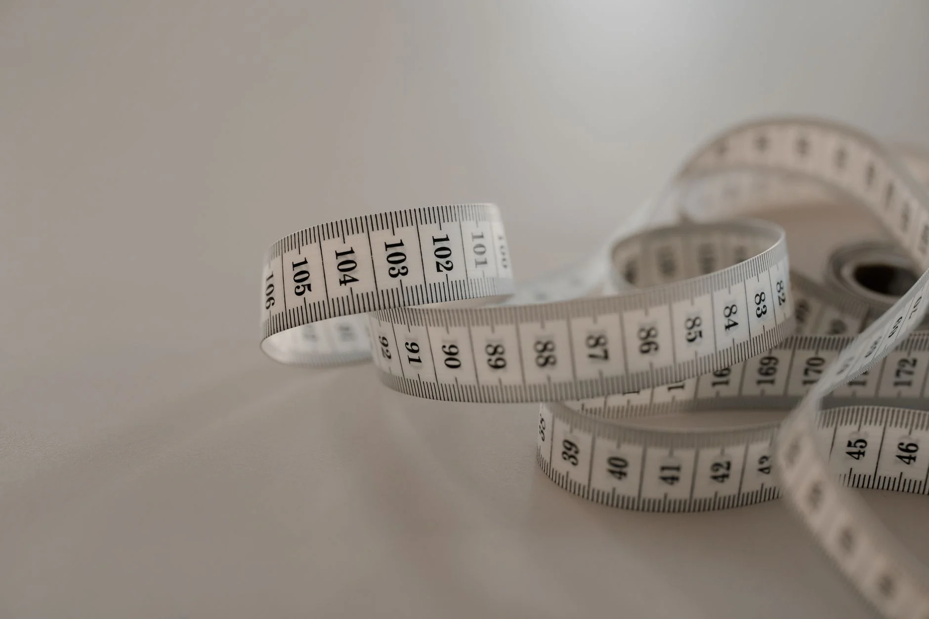 White tape measure with black numbers, coiled loosely on a plain surface.