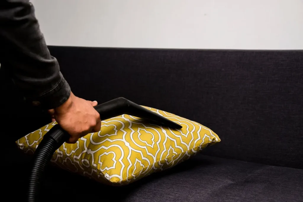 Close-up of a person vacuuming a patterned pillow on a dark sofa indoors.